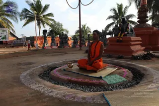 A photograph of an outdoor courtyard with sandy ground and palm trees. In the centre, a seated figure in orange robes and a rudraksha mala sits cross-legged on a woven mat atop a raised circular platform of dark gravel, which is bordered by white and pink rangoli-style patterns. A small brass vessel sits beside the seated figure. Behind, rows of dark stone deity murti and figures stand on red bases, and a large black stone sculpture with garlands occupies the right. A red pillar and a red brick wall are visible, along with a signboard at the left. The seated figure is a person with a shaven head, not a statue, so the a person cue is orange robes with a rudraksha mala while seated in meditation.