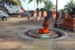A photograph of an outdoor ashram courtyard on bare earth. In the centre, a seated man in orange robes sits cross-legged on a mat and cushion inside a circular stone-edged pit lined with dark gravel and a pink-and-white ring, his hands pressed together in prayer, with a small brass vessel beside him. A thin man with a shaved head and a sleeve-less orange wrap, a rudraksha-style cord at his neck, meditates. Behind him stand several dark stone statues on red pedestals, including seated figures and a standing deity, and a red-orange wall. A woman in a pale yellow sari walks in the background, and a silver pickup truck sits at left. A large map-like board with coloured diagrams hangs under a thatched roof, and a garland of orange and white flowers drapes a statue at right.