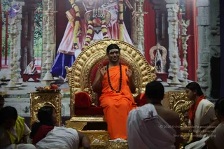 A man in saffron-orange robes and a dark rudraksha mala sits on an ornate gold throne with a red cushion, hands raised in a gesture while speaking. He has a sandalwood or vibhuti mark on his forehead and long swept-back hair, with a shawl draped over his shoulders. Around him, devotees sit on the floor in white dhotis and white garments, and a woman in a white sari with a red-bordered shawl sits at right. Behind the throne is a large printed backdrop of a temple scene with a deity figure garlanded in yellow and red flowers, brass lamps and carved stone pillars, plus a fruit offering on a gold side table. The scene suggests a satsang discourse.