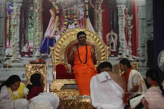 A man in an orange saffron robe and a dark rudraksha mala sits on an ornate gold throne with red cushions, raising both open hands in a teaching gesture. He has a red tilak on his forehead. In front of the throne, several people in white ritual clothes with gold borders sit on the floor, including a woman in a yellow shawl on the left and a woman in an orange shawl on the right. A gold-framed table holds fruit and offerings. Behind the throne is a large backdrop of a deity with garlands, and gold lamps stand on both sides. The a person candidate shows saffron robe, rudraksha mala and a golden throne, a probable match. Seated devotees sit near the throne during satsang.