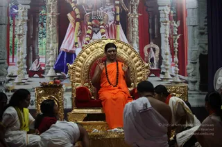 A man in a saffron-orange robe and a dark rudraksha mala sits on an ornate gold throne with a red cushion, hands raised in a blessing gesture, while devotees in white dhotis and saris with yellow and red borders sit around and before him. Behind the throne a large painted backdrop shows a deity figure with garlands and a red-lined cloth, flanked by tall brass lamps. A fruit offering and small silver items rest on a gold stand in front, and a fan is visible at the right edge. Several people are visible in the foreground, seated or bending forward.