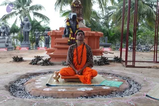 A man in saffron-orange robes sits cross-legged in meditation with eyes closed and hands folded in his lap, on a raised circular earthen platform ringed with gravel and decorated with white rangoli dots and pink line motifs. He wears a leopard-print shawl over his shoulders and a dark rudraksha mala around his neck. Beside him stands a brass pot on the matting. Behind him, a dark stone Shiva lingam wrapped in a garland of yellow and orange flowers sits on a red-orange pedestal. Grey stone deity statues, palm trees and red placards stand in the garden. a person: the saffron robes, long swept hair, rudraksha mala and leopard shawl are distinctive visible cues for the presenter, shown in a meditation posture.