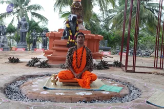 A man sits cross-legged in meditation on a raised circular earthen platform ringed with gravel and decorated with white floral rangoli, eyes closed, hands resting in his lap. He wears an orange-saffron dhoti and shawl over a leopard-print top, with a dark rudraksha mala around his neck. Behind him stands a dark stone Nandi bull with a garland of marigolds and red flowers, set on an orange-red tiered pedestal. Around the scene are dark grey stone deity statues, a brass kalasha-style vessel sits beside him on a woven mat, and palm trees and red metal railings frame the outdoor ashram grounds.