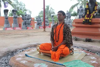 A man sits cross-legged in meditation on a raised stone platform outdoors, eyes closed and hands resting in his lap. He wears saffron-orange robes under a leopard-print shawl and a long dark rudraksha mala, with a red tilak on his forehead. A brass vessel and a woven mat sit beside him, and white kolam-style floral designs decorate the ground. Behind him stand grey stone sculptures of deities and sages, a red-and-white board with text, and an orange pedestal draped with a marigold garland. Palm trees and an orange wall complete the setting. A seated meditator fits the page's meditation theme; his identity is not established from appearance, as the cues are limited to saffron clothing and a mala.