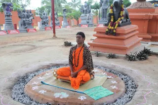 A seated man in an orange saffron robe and a leopard-print shawl sits cross-legged in meditation on a raised circular platform outdoors. He wears a dark rudraksha mala and holds his hands in his lap, eyes closed. Beneath him is a woven mat with a blue-edged cloth and a brass vessel. The circle is ringed with grey gravel and decorated with white rangoli motifs. Behind him stands an orange-red stepped pedestal topped by a black Nandi bull statue draped in a yellow and red flower garland. Grey stone deity sculptures and a row of red placards line the courtyard. This appears to be a person figure, with the robe, leopard shawl and rudraksha mala as qualifying cues, shown meditating in a temple-style courtyard.