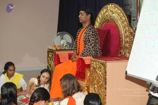 A satsang photograph in a hall. A man with long swept-back dark hair sits cross-legged in meditation on an ornate gold throne with red cushions, wearing an orange saffron dhoti and a leopard-print top, with a long orange-and-black beaded mala around his neck; he is a probable Swamiji by these cues. Seated on the floor in front, a woman in a yellow-bordered white sari and a woman in a cream sari with hair down attend to brass plates containing red flowers. Other seated attendees in orange and white clothing are visible in the foreground. A white board with handwritten diagrams stands at right, a pedestal table holds apples and flowers, and a fan sits behind.