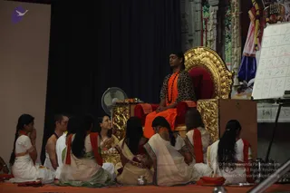 A seated man in an orange saffron garment and a leopard-print top sits on an ornate gold throne with a red cushion, wearing a long rudraksha mala. Several devotees sit cross-legged on an orange cloth before him: a woman in white with a folded-hands namaskar, a woman in a cream sari with a red border, and others in white and cream garments with red shawls. A small brass pot and offerings sit near the throne, a whiteboard with letter grids stands at right, and a white pedestal fan is at left. The person on the throne has distinctive long hair, a rudraksha mala and saffron clothing, and is a person candidate in an appearance-based label. Around twelve people are visible.