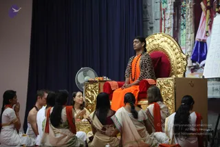 A seated man on an ornate gold-and-red throne, wearing an orange dhoti and a leopard-print top with a rudraksha mala, sits before a seated group of devotees. Several women in cream and white saris with orange-red borders sit on the floor with hands folded in namaste and one holds a flower, while a man in white and a woman in white sit nearby. A small table holds fruit and offerings, and a white fan stands at left.