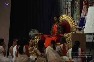 A seated man in an orange saffron dhoti and a leopard-print top sits on an ornate gold throne with red cushions, wearing a dark rudraksha mala with orange beads, and holds his hands together in front of him. Nearby stands a small table with a fruit offering and a small lamp. Around the throne sit devotees in white and cream sarees and dhotis with red and orange shawls, several with hands folded in anjali mudra, including a woman in a white sari with a striped border. A white standing fan and a whiteboard showing a grid of letters stand to the right, with a dark curtain and a colourful deity-decorated backdrop behind. a person: the seated man wears saffron, rudraksha beads and sits on a gold throne, consistent with the satsang setting.