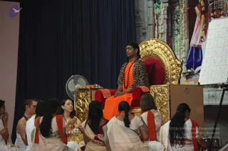 A photograph of a satsang hall where a man seated on an ornate golden throne with red cushions wears a leopard-print jacket over an orange garment, a rudraksha mala and a long swept-back hairstyle, matching the page's a person cues. Several devotees in white and cream sarees and dhotis with orange shawls sit on the floor before him, hands folded in namaste or resting in their laps, with a brass offering plate holding fruit and flowers beside the throne. A standing fan and a navy curtain stand behind the stage, and a whiteboard with a grid of letters is at right.