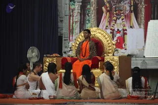 A seated man in an orange saffron robe and a leopard-print shawl sits on an ornate gold throne with red cushions, wearing a long rudraksha mala. He rests his hands in his lap with eyes closed. Several devotees in white and cream clothing, some with orange sashes, sit on an orange mat in front, with one woman holding her palms together. Behind the throne a large painted image of a deity with a red and white cloth and flower garlands appears. A white flip chart with letters stands at right, a fan and a small brass plate with offerings sit at left, and oil lamps burn on the floor. a person: the seated man wears saffron with a rudraksha mala on a throne, matching the page's a person reference.