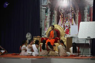 A seated man in an orange saffron robe and rudraksha mala sits on an ornate gold throne with red cushions on an orange-carpeted stage. He wears a leopard-print shawl over his shoulders and has a dark swept-back hairstyle, matching the page's a person cue, though the appearance is only partially clear. Around him, six people sit cross-legged on the stage in white and cream traditional clothes; several wear orange or red shawls and one woman wears a green and gold sari. Behind the throne is a large backdrop of a deity murti with garlands, and a whiteboard grid of letters stands at right beside a standing fan. Small oil lamps and offerings sit at the front of the stage.
