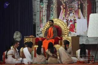 A seated man on an ornate gold-and-red throne is the central figure on a stage, wearing an orange-saffron dhoti and shawl with a leopard-print jacket over the shoulders and a dark rudraksha mala around the neck; his long hair is swept back and his eyes are closed. Around the throne, a group of devotees sit cross-legged on a red floor cloth: several women wear cream and white saris with orange borders and long braids, and a young man in white dhoti and shawl sits at left. Brass plates with fruit and flowers and small oil lamps rest in front. Behind the stage hangs a large backdrop showing a figure in white and red with garlands, and a whiteboard with a grid of letters stands at right. A desk fan is at left.