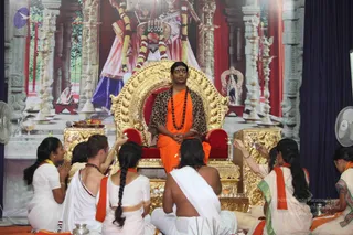 A seated man on an ornate golden throne with red cushions at the front of a ceremonial hall, wearing an orange saffron wrap, a leopard-print shawl over his shoulders, and a long rudraksha mala with a red-dotted forehead mark. a person is the candidate for this seated figure based on the orange robe, rudraksha mala and ornate setting. Around the throne, several devotees in white garments with orange shawls sit on the floor, some with palms joined and others holding small brass vessels and offering items. Behind the throne is a large backdrop of a deity murti with pillars, brass lamps and a red curtain, and the scene appears to be a satsang or puja gathering.