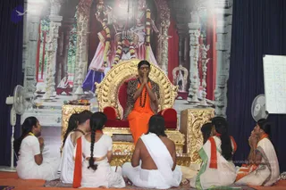 A satsang in a temple-style hall. A man seated on an ornate gold throne with red cushions, in an orange robe, rudraksha mala and leopard-print shawl, sits with palms pressed together in namaskar. A backdrop shows a painted temple scene with a deity murti and garlands. Around him, six devotees sit on a patterned carpet: women in white and cream clothes with orange shawls, and a man with bare upper body in white dhoti. Two oil lamps and a small tray of offerings sit on a gold stand beside the throne. Fans stand at the sides and a whiteboard with letters is on the right.