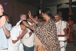 Photograph of a satsang-style greeting indoors. A central figure with long dark hair tied back wears a leopard-print shawl over orange clothing and raises a hand in blessing, placing his palm on the head of a bowing man in white shirt and white trousers with a rudraksha mala. Nearby, a man in a light blue shirt presses palms together, and other men in white shirts, a checked shirt and glasses stand behind. On the right, a shaven-headed young man in white dhoti with a gold chain and a tilak holds a white box with red trim. Saffron-robed men stand further back. Colourful striped pillars and white drapery decorate the hall.