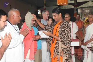 A photograph of an indoor ashram gathering. A man with long dark hair, orange robe, rudraksha mala and a leopard-print shawl stands center-right, placing his hand on the head of an elderly man wearing a grey knit cap, glasses and a salmon-pink shawl, who holds folded hands and a white envelope. Several people in white clothes watch with folded hands, including a bald man in white with a shawl at left. Other shaved-headed men in white dhotis and orange cloth with white forehead markings stand at right, one holding a small red-and-white box. A decorated backdrop with a framed picture and orange draping fills the background.