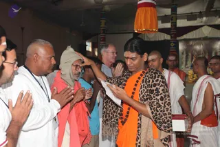 A photograph of a temple-hall ritual in which a long-haired man in an orange saffron cloth and a leopard-print shawl with a dark rudraksha mala stands centre frame, pressing his palm to the forehead of an elderly man in a grey knit cap and pink-orange wrap who holds his hands in anjali. Around them, a man in a white shirt with a lanyard holds his hands together, a younger man in a blue shirt stands nearby, and temple priests in white dhotis with red borders and bald-shaved heads stand at right. The orange-and-white banner hangs from the pillar above, with colourful painted columns and a shrine decorated with deity images behind. The scene reads as a blessing or initiation moment.