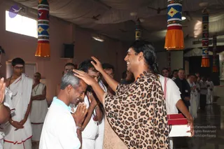 A photograph of an indoor hall with pink walls, white ceiling drapery and hanging striped banners with orange tassels. In the foreground a man in a bright blue collared shirt and a white dhoti-style garment stands with his head bowed and hands folded in namaskar, while a long-haired person in a leopard-print shawl and a white garment places a palm on his head in blessing. Around them, people in white ritual clothes, some with white tilaka marks, stand and watch. Two red boxes are held at the lower right. A probable Swamiji candidate with long swept-back hair, orange garland and rudraksha beads gives the blessing.