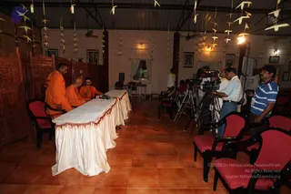 A photograph of a press meet in a hall with terracotta tiled floor and wooden carved screens at left. Three men in saffron-orange robes sit behind a long table draped in white fabric with a red fringe, with a microphone on the table. A bald man in saffron stands at the left edge of the table. On the right, several cameramen with video cameras on tripods film the proceedings, and a man in a blue striped shirt stands near them. Rows of red-cushioned chairs face the table. Paper crane decorations hang from the ceiling, and framed portraits and pictures decorate the walls.
