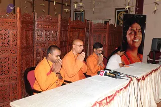 A press meet at a wooden-screened hall. Three men in saffron-orange robes sit behind a draped white table with red-trimmed edging, palms pressed together in namaste; the man on the right wears wire-rimmed glasses and a rudraksha mala, and the man in the centre is bald and wears glasses. Microphones and papers sit on the table. A woman in a white top sits at the far right end writing. Behind them stands a large portrait of a smiling woman with a red bindi and orange garment, and small framed deity images hang on the wall. Paper crane garlands hang from the ceiling.