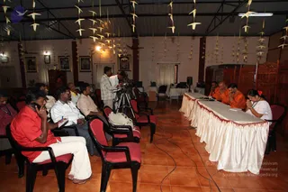 A press meet in a hall with a terracotta tiled floor and paper cranes hanging from the ceiling. At the right, a long table draped in white cloth with a red fringe seats several saffron-robed men with shaved heads and a woman in white with hands pressed in namaste. Seated audience members face the table, including a man in a red shirt and white trousers talking on a mobile phone and another in a white shirt taking notes. A cameraman films from a tripod near the middle, and framed devotional pictures hang on the walls. Several chairs with red cushions are arranged in rows. Visible people number roughly 15-20.
