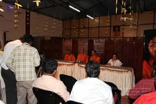A press meet in a wooden-roofed hall decorated with hanging paper cranes. At a long table draped in cream cloth with a red trim, three men in saffron-orange robes sit beside a woman in a white sari with a dark shawl. Microphones sit on the table. Behind them, carved wooden panels form a screen. A framed portrait of a woman in orange hangs on the wall, and a large banner of a smiling person in orange stands at the right edge. In the foreground, seated audience members in white and checked shirts face the table, and a cameraman stands at left with a video camera on a tripod.