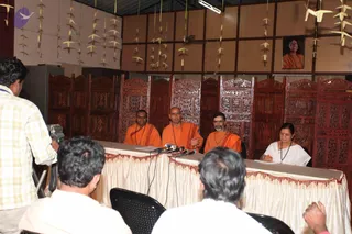 A press conference in a wooden-panelled hall with carved screens and paper crane decorations hanging from the ceiling. Three robed men sit behind a draped white table with red fringe: two on the left in saffron-orange robes, and a bald man in the centre in orange robes gesturing as he speaks. A woman in a white sari with a dark beaded necklace sits at the far right writing on paper. Microphones sit on the table. A framed portrait of a woman in orange hangs on the wall. A cameraman in a yellow-checked shirt films at left, and seated journalists are visible in the foreground. The centre man has a shaved head and plain orange robes with no long hair, crown or rudraksha cues, so his identity is uncertain.