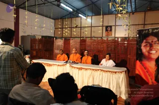 A press conference in a hall with carved wooden screens and a cream-skirted table draped in gold trim. Three saffron-robed men sit behind the table; the centre one has a shaved head and the others are bare-headed, with beaded necklaces. A woman in white sits at the right end writing. Behind the table hang yellow paper garlands and a framed portrait of a smiling woman with a red bindi. A photographer stands at left and several seated audience members in the foreground face the table. A large banner at right shows a smiling woman in orange with dark hair, not clearly the same person as the seated men. a person candidate appearance is uncertain, so the identity is left unsure.