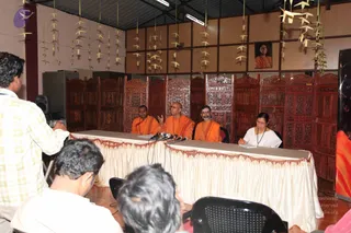 A press conference in a wooden-panelled hall with carved dark-wood screens and paper garlands hanging from the ceiling. Three men in saffron-orange robes sit behind a draped white table with microphones; the centre man, bald and gesturing with one hand, speaks. A woman in a white sari with a dark shawl and glasses writes on a pad at the right end of the table. A framed portrait of a woman in orange hangs on the wall behind them. In the foreground, a man in a yellow checked shirt holds a camera, and two seated audience members are partly visible.
