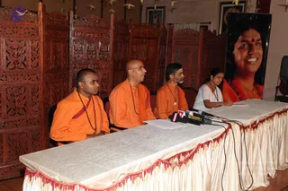 A press conference at a table draped in cream cloth with a red fringe, with several microphones on the table. Three men in saffron-orange robes sit behind it: the one on the left wears a red shawl and a rudraksha mala, the middle one is bald with glasses and a mala, and the one on the right has a grey beard and wears a mala and a pendant. A woman in a white top and glasses sits at the right, writing on papers. Behind them stands a carved wooden room divider and a tall vertical banner of a smiling woman with a red bindi and a garland, beside framed pictures on a wall.
