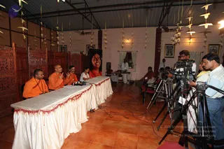 A press meet in a hall with terracotta tiled floor and wooden carved partitions on the left. Three men in saffron-orange robes sit behind a long table draped in white cloth with red fringe; the man in the centre, with a shaved head, gestures with both hands while speaking. A woman in an orange sari sits at the table's end. Behind them hangs a large portrait of a smiling woman in orange. Several cameras on tripods film from the right, with camera operators in casual clothes. Other people sit on red chairs at the back, and a man in white stands near the rear. Paper stars hang from the ceiling, and framed deity paintings decorate the walls. Nine or so people are visible, including partial figures at the edges.