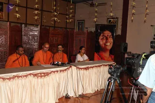 A press conference table draped in cream fabric with red fringe, with microphones on it. Four people sit behind the table: three men in orange saffron robes with rudraksha malas, and a woman in white on the far right with a mala and papers. A large framed portrait of a smiling figure with long dark hair and a red bindi, wearing orange, stands to the right behind the table. Carved wooden dividers, paper garlands and framed pictures decorate the room. A camera on a tripod and a video camera film the event from the right, and a cameraman stands at the edge.