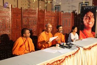 A press conference at a seated panel behind a draped table with several microphones and papers. Four people sit in front of carved wooden room dividers: on the left a young man in orange robes and rudraksha mala, in the centre a shaved-headed man in orange robes with a red tilak holding a sheet of paper and gesturing, beside him a bearded man in orange robes and mala, and at the right end a woman in a white top with glasses holding papers. On the right stands a large banner of a smiling person with long dark hair in orange clothing and a red bindi, with a rudraksha mala. Paper streamers hang from the ceiling in the background.