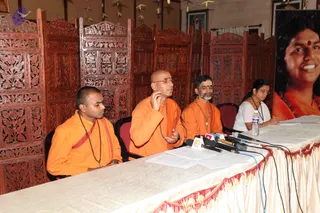 A press conference at a long table draped with a cream cloth edged in red fringe, with several microphones and papers on top. Four people sit behind it in front of an ornately carved wooden screen. On the left, a young shaved-headed man in orange robes and a red shawl with rudraksha beads sits with eyes lowered. Beside him, a bald man in orange robes and wire-rimmed glasses gestures with one hand while speaking. Next to him, a bearded man in orange robes and a pendant necklace sits with hands clasped. At the far right, a woman in a white top and dark beaded necklace reads papers. On the right, a large framed portrait of a smiling woman with a red bindi and orange garment is displayed, labelled with no readable text.