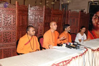 A press conference in an indoor hall with carved dark wooden screens behind a long table draped in a silver cloth with red fringe. Four people sit at the table: on the left a young man in orange robes with a rudraksha mala and a red shawl; next to him a bald man in orange with glasses and a mala, gesturing with one hand while speaking over a bundle of microphones; a bearded man in orange with a mala and pendant; and at the far right a woman in a white sari with glasses and a red bindi, with a water bottle and papers on the table. On the right edge a large banner shows a smiling woman in orange with a beaded necklace. Hanging paper decorations are visible at the top.