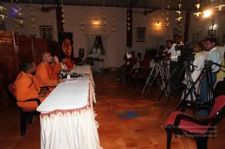 A press meet in a hall with terracotta tiled floor and paper bird decorations hanging from the ceiling. Two saffron-robed men sit at a long white draped table at left; the man nearer the table has a shaved head and gestures with palms pressed together, and the other man beside him wears a saffron robe and a rudraksha mala. A woman in white sits near the table. On the right, several cameramen and camera operators stand behind tripods and video cameras while others sit in red chairs facing the table. A large portrait of a smiling woman hangs on the back wall beside framed religious pictures and wooden carved screens at left.