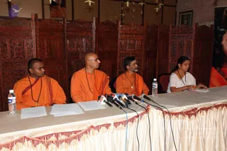 A press conference table draped in cream fabric with a red and gold fringe, with four people seated behind a row of microphones. Three men on the left wear saffron-orange robes and rudraksha malas, and the man on the far left wears a red-and-orange sash. The woman on the right wears a white sari and a mala, and holds a pen over papers. Water bottles and loose printed sheets sit on the table. Behind them are carved dark wood screens and a framed portrait on the wall. The image shows a press meet, with no identifiable a person depiction confirmed by visible cues beyond the robes and malas.