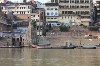 A photograph taken from the Ganges river looking toward the riverfront at Varanasi. In the foreground, several wooden boats are moored on the brown water. At left stands a tall stone shrine with a stepped, spired roof, partially submerged at the water's edge, beside a smaller stone structure. Behind them rise multi-storey buildings with balconies and shuttered windows, including a building with a blue sign reading Scindia Guest House. Stone ghat steps lead down to the river at right, where a few people are visible near the water and along the steps. Small figures can be seen sitting at the top of the steps at left.