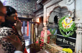 A photograph inside a stone shrine with a carved white marble niche. On the right, a dark cloth-draped murti stands adorned with a large green leaf garland, orange and white flowers and a yellow marigold garland, with Devanagari lettering on the black cloth reading Kaal Bhairav. In the centre a brass oil lamp with a lit flame stands on a tall stand. A man in a red shirt and red dhoti stands behind a brass railing near the murti. In the foreground at left, a person in a leopard-print top with palms pressed together in namaskar, wearing a light tilak, stands in devotion. Yellow marigolds lie on the lower right and a brass pot sits near the lamp. The walls are tiled in green and red.