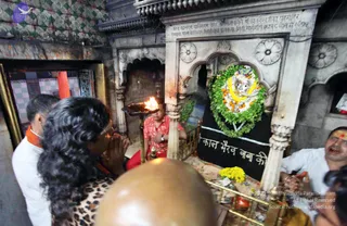 A photo inside a small shrine chamber where a priest in a red-and-pink checked shirt holds a flaming oil lamp (aarti) before a decorated stone deity murti with a face-like carved image wreathed in green leaves, marigolds and white flowers. Yellow marigold offerings sit on the floor and a black cloth below reads 'काल भैरव बाबा की'. On the left a woman with long curly black hair and a leopard-print top stands with palms pressed together in namaste, beside a man in white with an orange shawl. On the right a man with a tilak on his forehead in a white shirt claps or gestures, and a shaved-headed person's bald head is in the foreground. Brass railings and marble pillars frame the scene. Several people are visible.