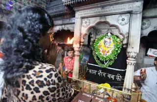 A photograph inside a stone shrine at a temple. In the foreground, a person with voluminous dark curly hair and a leopard-print top stands with palms pressed together in prayer, facing a small shrine. Behind them a man in a red patterned shirt with a necklace stands beside a brass railing. A flame burns in a brass lamp at left. The shrine's central niche holds a silver-faced deity murti garlanded with green leaves and orange and white flowers, draped in a black cloth with white lettering, and a yellow marigold garland lies below. A brass donation box sits in the foreground. A man in white with red and white wrist threads stands at right with hands raised.