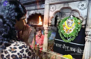 Photograph inside a marble shrine at a temple. In the foreground, a woman with dark braided hair and a leopard-print top stands with palms pressed together in prayer, facing a tall brass oil lamp whose flame burns brightly. Behind the lamp, a man in a red-and-white patterned shirt sits on a red mat, looking toward the camera. To the right, a silver face-mask murti of a deity is adorned with a green leaf garland, yellow and orange marigold garlands and white flowers, set before a black cloth with a white Hindi inscription. Carved marble arches and floral reliefs frame the shrine, with yellow flowers in the lower right.