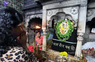Photograph inside a stone shrine with carved white marble arches and an inscribed plaque above. A woman with curly dark hair and a leopard-print top, seen from behind, holds her hands in anjali near a brass oil lamp with a lit flame. In the centre, a seated priest in a red-and-white patterned shirt and red cloth sits beside a silver-faced deity murti garlanded with green leaves, white flowers and orange marigolds, backed by a black cloth with Hindi lettering. A white-clad devotee with folded hands is at the right edge, and yellow marigolds lie at the base of the shrine. Brass railings frame the scene.