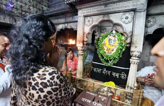A photograph inside a temple shrine. In the foreground a person with long dark curly hair and a leopard-print top stands with palms pressed together in prayer, facing a white marble shrine. Beside them on the left, a man with a tilak mark on his forehead wears white and orange. Behind the shrine, a man in a red patterned shirt sits near a brass lamp with a burning flame. The shrine holds a deity murti decorated with a green leaf garland, orange and white flowers, and a yellow marigold cloth, set in front of a black cloth with white Hindi lettering. A brass donation box with Hindi script sits at the bottom, and a yellow flower offering lies on the floor. Several other people in white stand at the right edge.