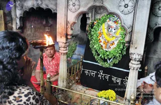 Inside a carved white marble shrine, a man in a red and white patterned shirt and red trousers kneels and holds a brass tray of burning camphor flame toward a silver-faced deity murti draped in a black cloth. The murti is adorned with a large green leaf garland, yellow marigold and white flower garlands, and an orange flower tilak. A woman with dark curly hair in a leopard-print top stands in the foreground with hands folded in prayer. Yellow marigold flowers lie on the floor in front, and brass railings frame the shrine. Hindi text on the black cloth reads Kaal Bhairav Baba.