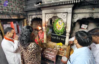 A photograph inside a small stone shrine with carved white marble arches and a Devanagari inscription above. A priest in a red shirt holds a lit aarti lamp with flames toward a deity image garlanded with green leaves and white and orange flowers, set against a black panel with Hindi lettering. In front, a woman in a leopard-print top and a man in a white shirt with an orange shawl stand with palms pressed together, while two men in light shirts look on from the right. A donation box marked in Hindi and English sits on the floor by yellow marigolds.