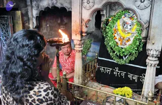 A photograph inside a stone shrine niche decorated with carved white marble arches and floral reliefs. A silver face of Kala Bhairava, a deity murti with a moustache and a tilak, is framed by a large green leaf garland and marigold and yellow flower garlands, in front of a black cloth with white Devanagari lettering. A man in a red shirt with white dots stands behind a brass railing, holding a flaming oil lamp on a tray. In the foreground a woman with dark wavy hair and a leopard-print top stands with her palms pressed together in prayer. A pile of yellow marigolds lies at the lower right.
