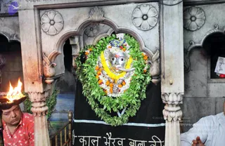 A photograph inside a shrine showing a silver face mask of a deity murti, adorned with a yellow and white marigold garland, orange flowers and green leaf garlands, set within a white marble arched niche with carved floral rosettes. Below the murti is a black cloth with silver border and Hindi lettering. On the left, a man in a red patterned shirt holds a flaming oil lamp, and on the right a partly visible man in a light blue shirt sits with folded hands.