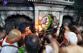 Crowded interior of a stone shrine in a temple. A carved marble niche holds a deity murti, a white stone figure decorated with a wreath of white and orange flowers, green leaves and a garland. A brass oil lamp with a bright flame sits in front. Around it, several shaved-headed priests in white dhotis and bare upper bodies, some wearing rudraksha malas and white sacred markings on the forehead, crowd close, reaching toward the shrine. A man in an orange cloth and white shirt stands at left with a red shawl. A carved inscription in Devanagari script runs across the top of the shrine wall.
